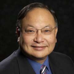 Dr. Ronald Mitsuyasu with short dark hair, wearing a dark suit, a blue shirt, and a striped tie. His expression is professional and friendly.