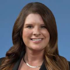 Emily Stimpson with long wavy hair, wearing a black blazer and silver necklace, posed confidently against a blue background.