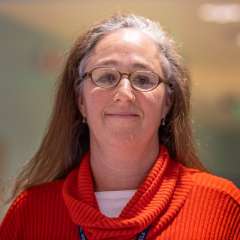 Gina Kornfeind wearing a red sweater, standing in a softly lit environment.