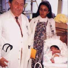 A vintage photo of two doctors, a man and a woman, standing next to a newborn baby lying in an incubator in a hospital setting.