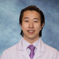 A headshot of Dr. Jerry Lee who has dark brown hair and is wearing a white lab coat, purple tie and smiling
