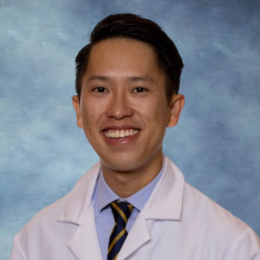 A headshot of Dr. Thang Tran who has short dark brown hair, wearing a white lab coat, tie and smiling