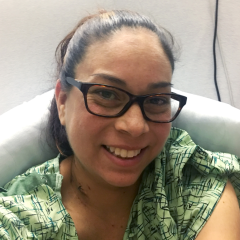  A portrait of a smiling Giorgiana in a hospital gown. She is wearing glasses and has her hair tied up in a ponytail.