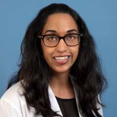 Headshot of Dr. Puja Venkat, with dark hair and glasses, smiling in a white lab coat. The pose implies she is friendly and professional.