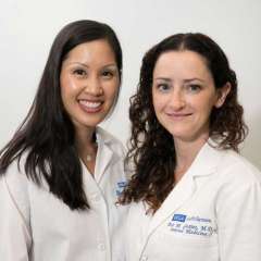 A portrait of two female medical professionals, one with straight hair and one with curly hair, both smiling and wearing white lab coats.