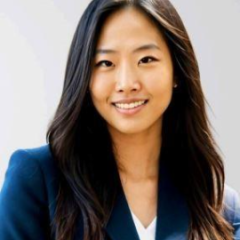 A woman in a dark blazer with long hair, standing against a light background.