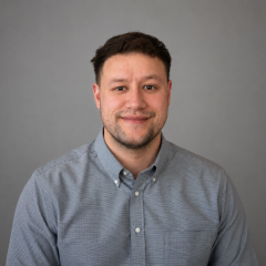 Daniel Cortez - Smiling man in gray button-down shirt against plain gray background.