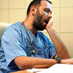 A male medical professional in blue scrubs, with a beard and stethoscope, yawns with his eyes closed and hand on his face.