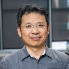 Headshot of Doctor Zhang wearing glasses and a grey button-down shirt, the background is blurry
