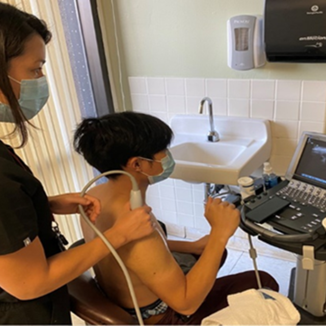 A woman with dark hair is performing an ultrasound procedure on a man's right shoulder.
